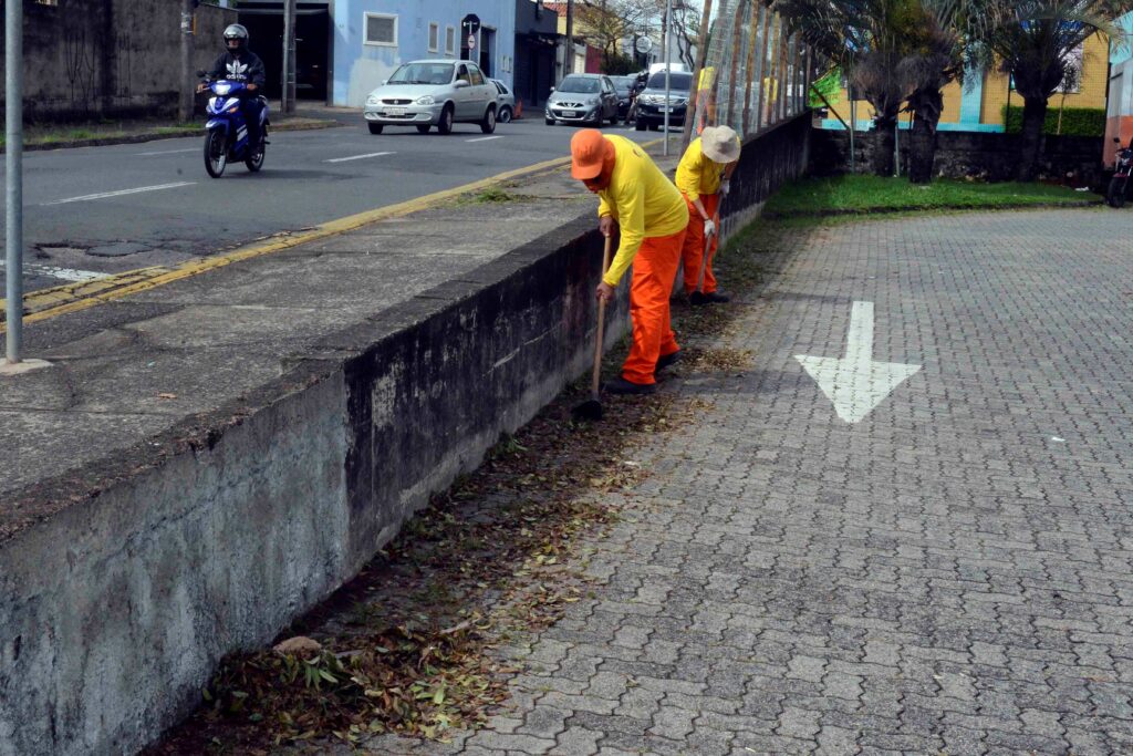 Dois trabalhadores equipados com fardamentos amarelos e laranjas utilizam enxadas para realizar a limpeza e capinação ao longo de um canteiro.