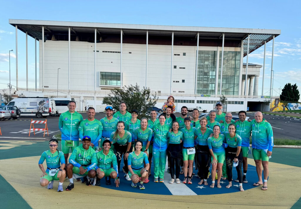 A equipa de triatlo e duatlo de Piracicaba posa reunida e a sorrir, todos vestidos com uniformes desportivos verdes e azuis correspondentes, em frente a um grande edifício moderno.