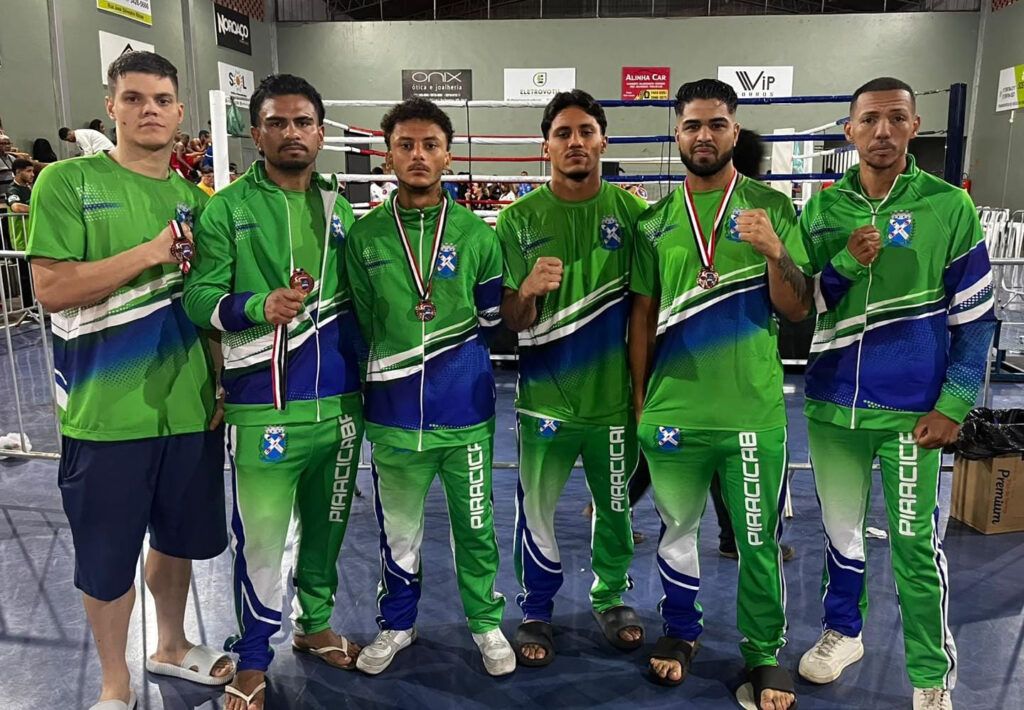 Seis atletas da equipa de boxe de Piracicaba vestindo agasalhos desportivos verdes e azuis da cidade posam lado a lado a exibir as suas medalhas de bronze dos Jogos Abertos em frente ao anel de boxe.