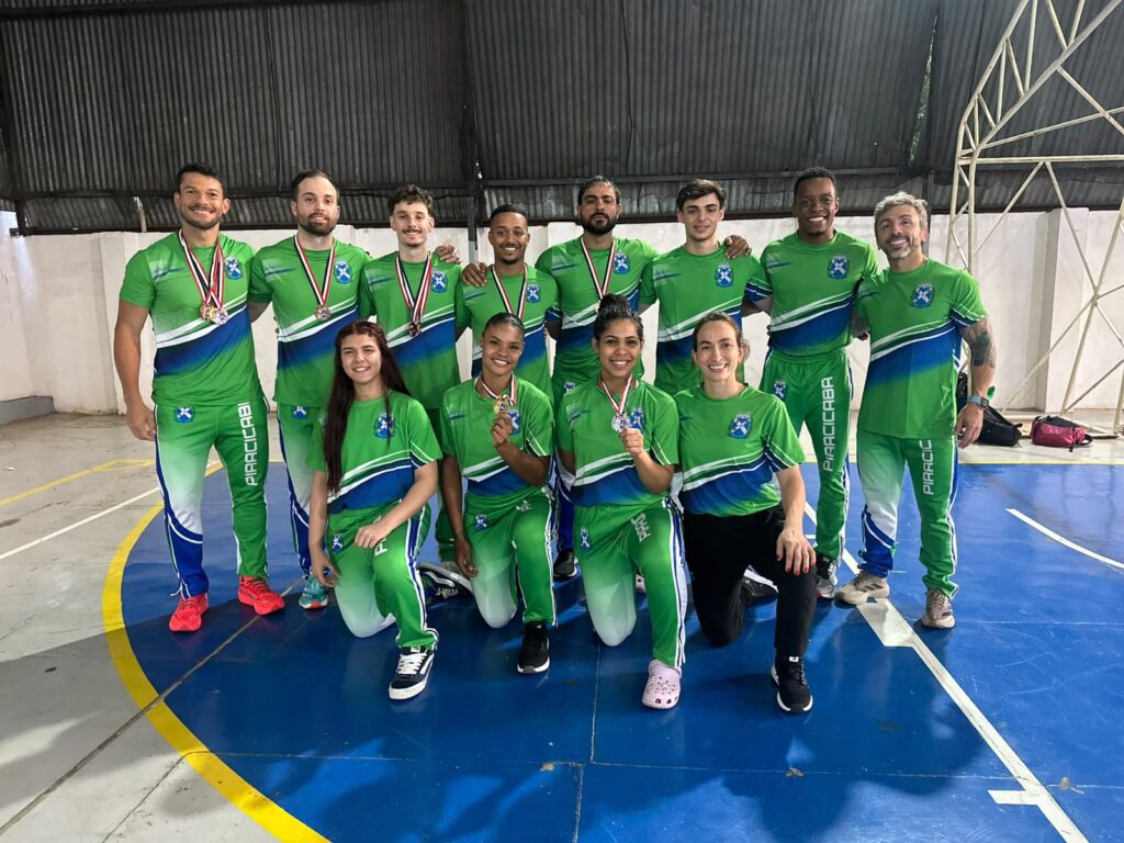 Equipa de karaté de Piracicaba reunida e posando num pavilhão desportivo com os uniformes da delegação, exibindo as medalhas conquistadas nos Jogos Abertos.
