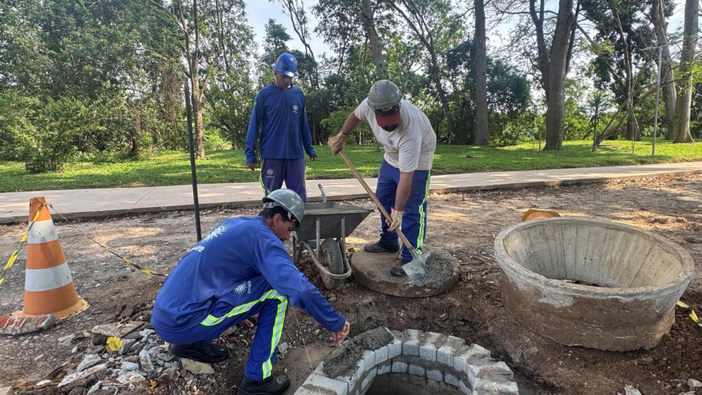 Três trabalhadores a aplicar massa e a ajustar a estrutura de alvenaria de um poço de visita numa área ajardinada.