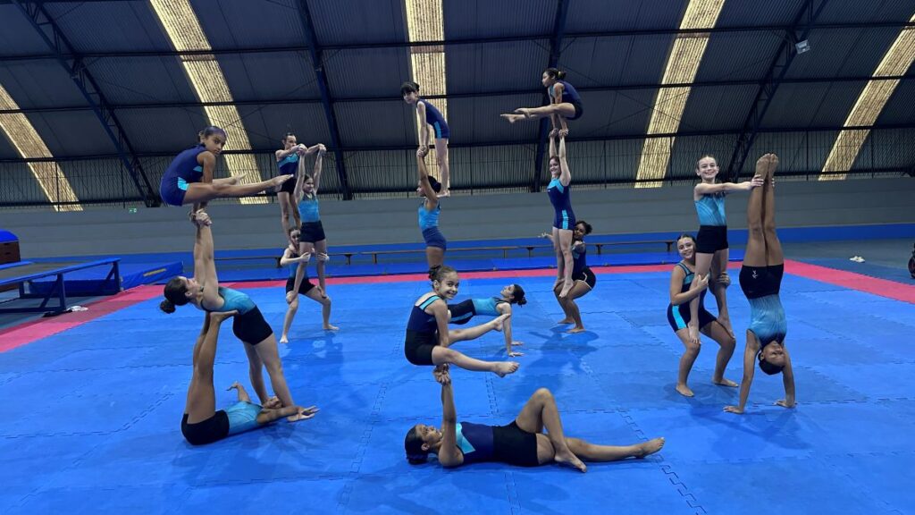 Integrantes da equipa realizam diversas pirâmides humanas e acrobacias sincronizadas sobre o piso azul do ginásio.