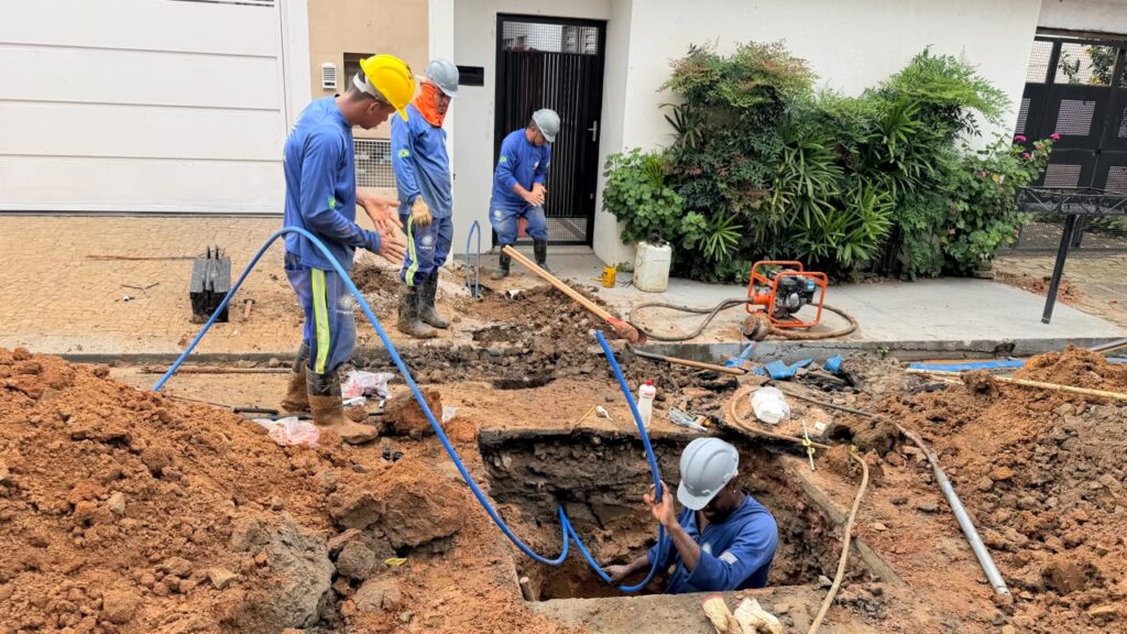 Mostra trabalhadores com capacetes e equipamento de segurança a manusear e a passar tubos azuis de água numa vala escavada na calçada, em frente a uma residência.