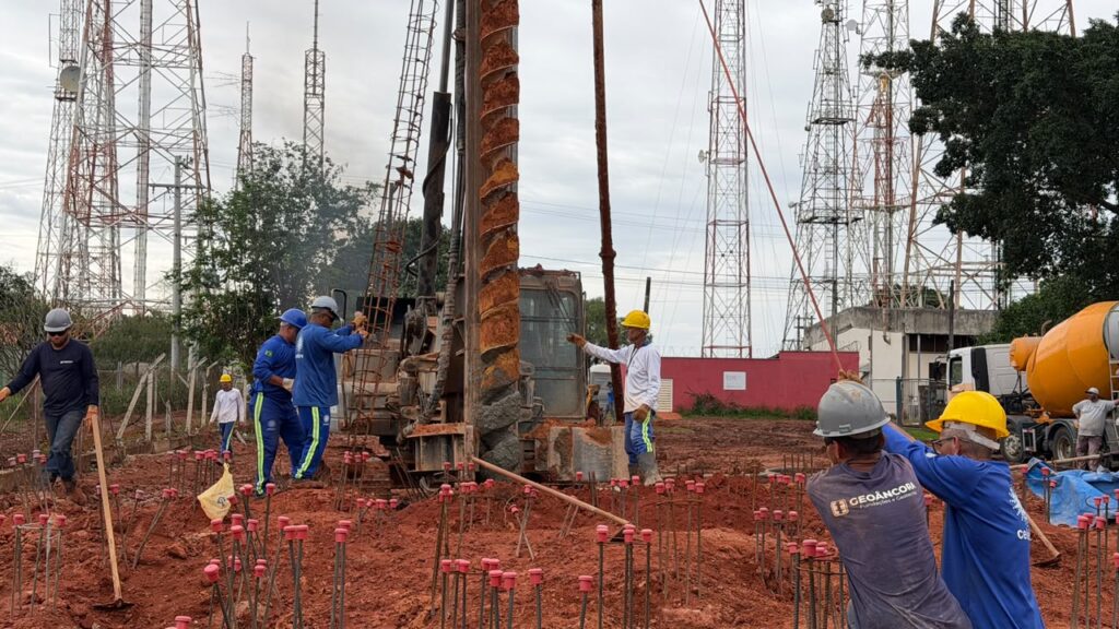 Operários com capacetes e vestuário de proteção trabalham no terreno de uma obra de fundação, onde uma perfuradora helicoidal escava o solo avermelhado repleto de estacas de aço, tendo várias torres de telecomunicações e um camião betoneira ao fundo.