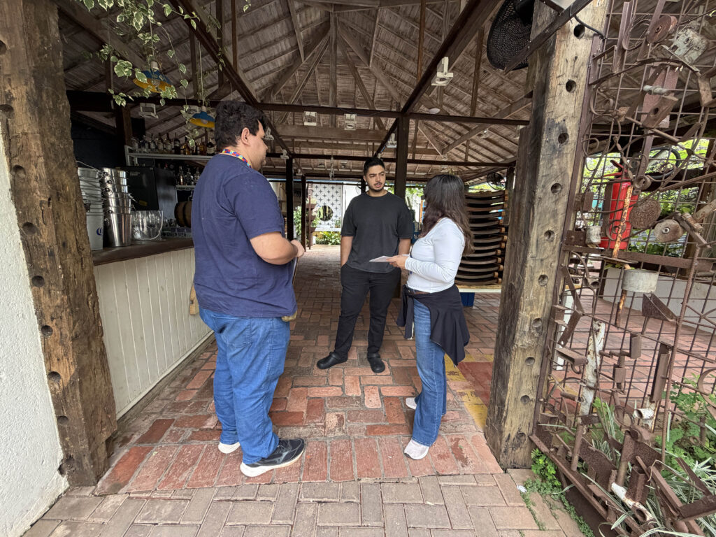 Três pessoas conversam no interior de um restaurante/comércio com estrutura de madeira e chão de tijolos; dois agentes prestam orientações a um funcionário/proprietário do local.