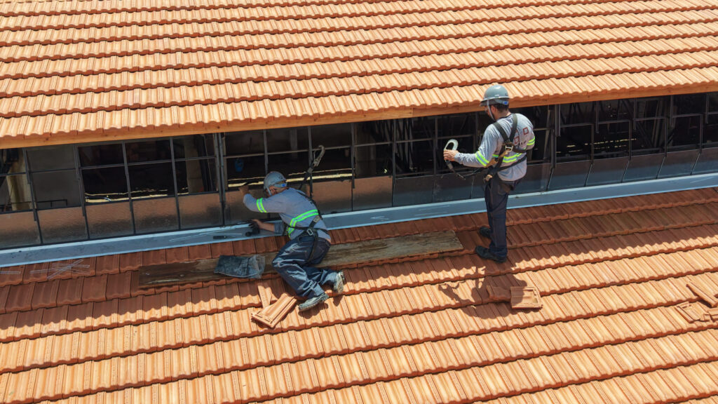 Dois operários com capacete e equipamento de proteção em cima do telhado de telhas cerâmicas a realizar a instalação de vidros nas janelas superiores.