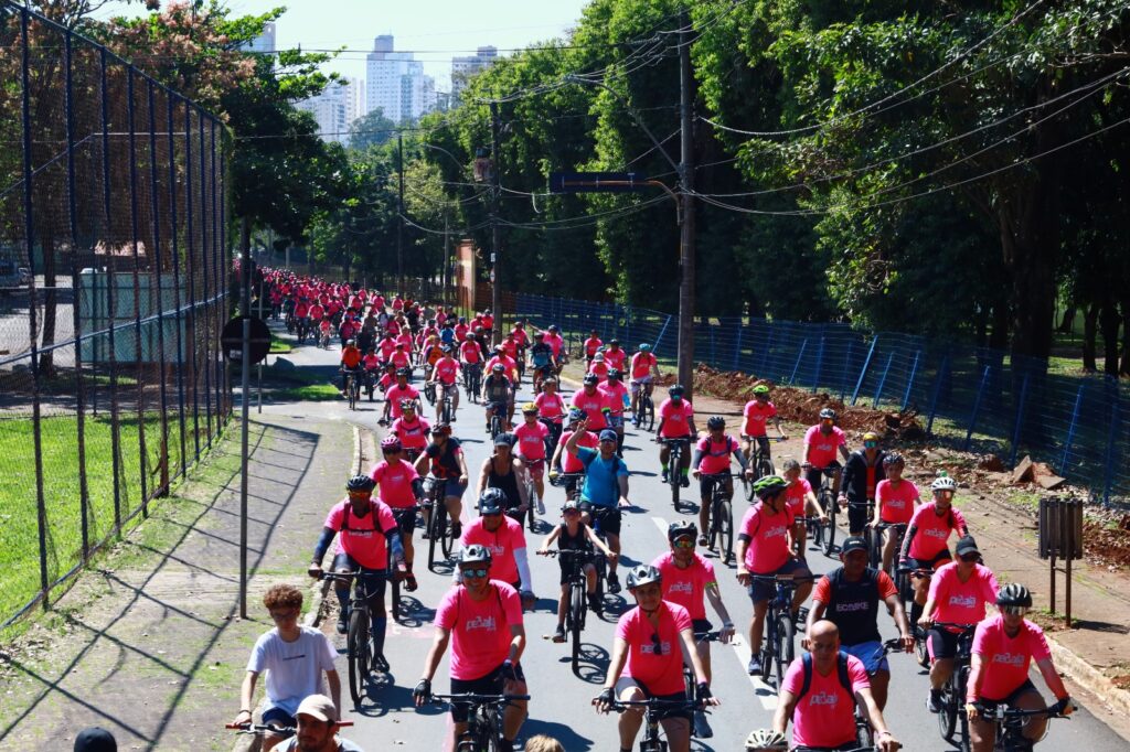Um grande grupo de ciclistas vestindo camisas cor-de-rosa pedala em conjunto por uma alameda arborizada durante o evento Pedala Pira.