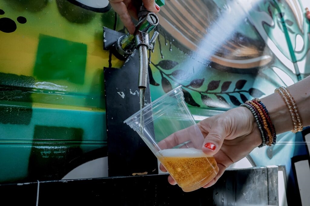 Um plano aproximado que mostra uma pessoa a segurar um copo de plástico transparente enquanto o enche com cerveja de uma torneira de chope acoplada a um veículo ou estrutura decorada.