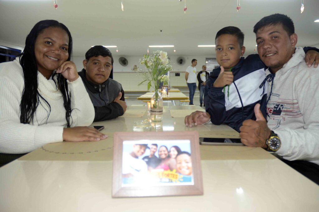 Integrantes da família Tabai sorriem sentados à mesa, onde há uma fotografia da família emoldurada, flores e uma vela acesa.
