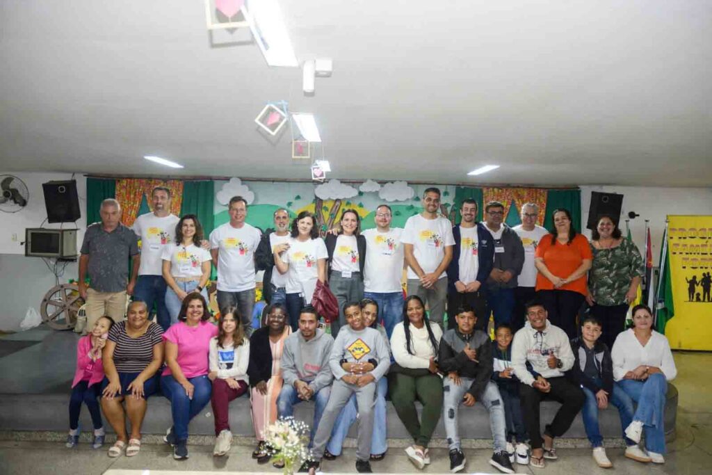 Grupo de adultos e crianças posa para uma foto coletiva no palco de uma escola durante a abertura do 2º ciclo do programa Famílias Fortes
