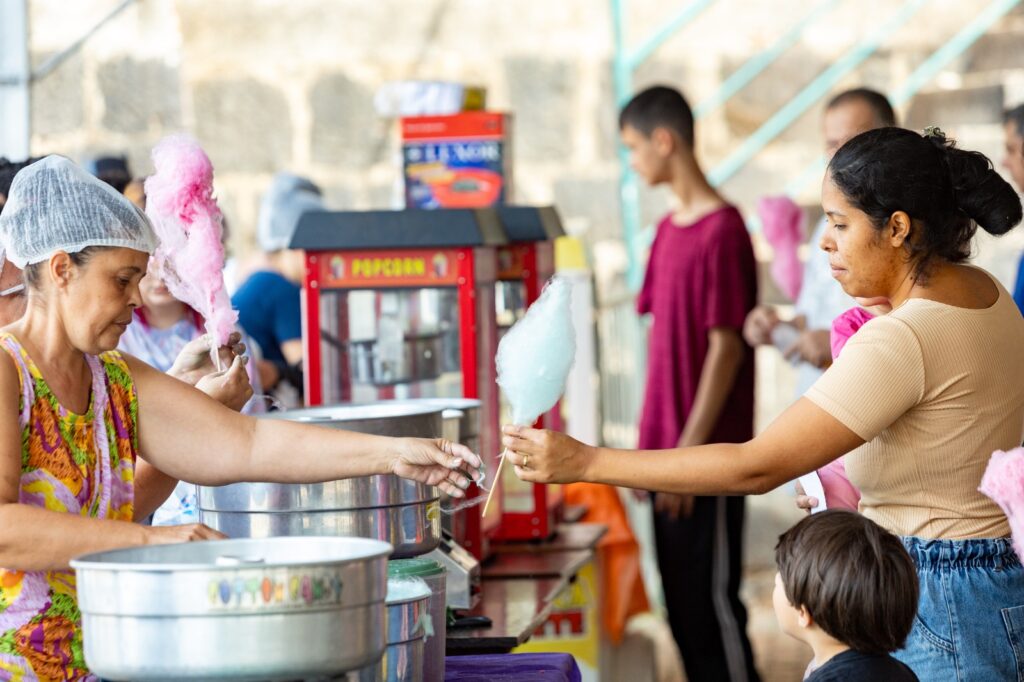Mulher a entregar algodão-doce azul a uma criança acompanhada pela mãe, numa banca com máquina de pipocas ao fundo durante evento comunitário.