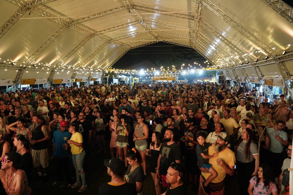 A multidão reunida sob uma tenda iluminada assiste a um evento noturno, possivelmente um show ou festival.

A imagem retrata uma vasta multidão de pessoas reunidas sob uma grande tenda com estrutura metálica aparente e um teto branco e texturizado, parcialmente iluminado por uma série de luzes de globo suspensas que criam um ambiente festivo e acolhedor. A iluminação principal vem de cima, projetando feixes de luz que criam sombras suaves no teto, além das cordas de luzes que pontilham o espaço, simulando um céu estrelado artificial. A cena é noturna, com a escuridão do céu visível em algumas aberturas da tenda e ao fundo. A multidão é densa, ocupando a maioria do centro e da frente da imagem, com pessoas em pé, observando algo fora do enquadramento, possivelmente um palco. Os indivíduos apresentam diversidade em termos de idade, etnia e vestimenta; muitos usam roupas casuais como camisetas, regatas e calças jeans. Há uma grande variedade de cores nas roupas, incluindo tons vibrantes e neutros, contribuindo para a atmosfera animada. Em algumas áreas da multidão, especialmente na frente, alguns rostos estão mais nítidos e focados, permitindo observar expressões de interesse e entretenimento. A iluminação da tenda, com tons quentes de amarelo e branco, emana uma atmosfera de celebração e união. A composição da imagem, tirada de um ângulo ligeiramente elevado, abrange uma grande área da tenda e do público, dando a sensação de imensidão do evento. Ao fundo, vislumbram-se mais luzes e possivelmente outras estruturas e barracas, indicando um evento maior, como um festival ou feira. A atmosfera geral é de excitação, alegria e comunidade, sugerindo um evento social bem-sucedido e apreciado pela multidão.