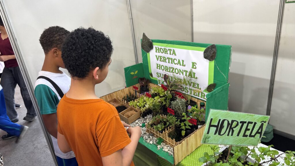Dois estudantes observam uma maquete interativa sobre horta vertical e sustentável acompanhada por vasos de hortelã num dos stands