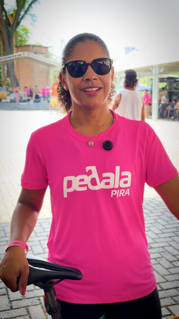Uma mulher sorridente usando óculos de sol e a camisa cor-de-rosa do evento Pedala Pira segura o selim de uma bicicleta.