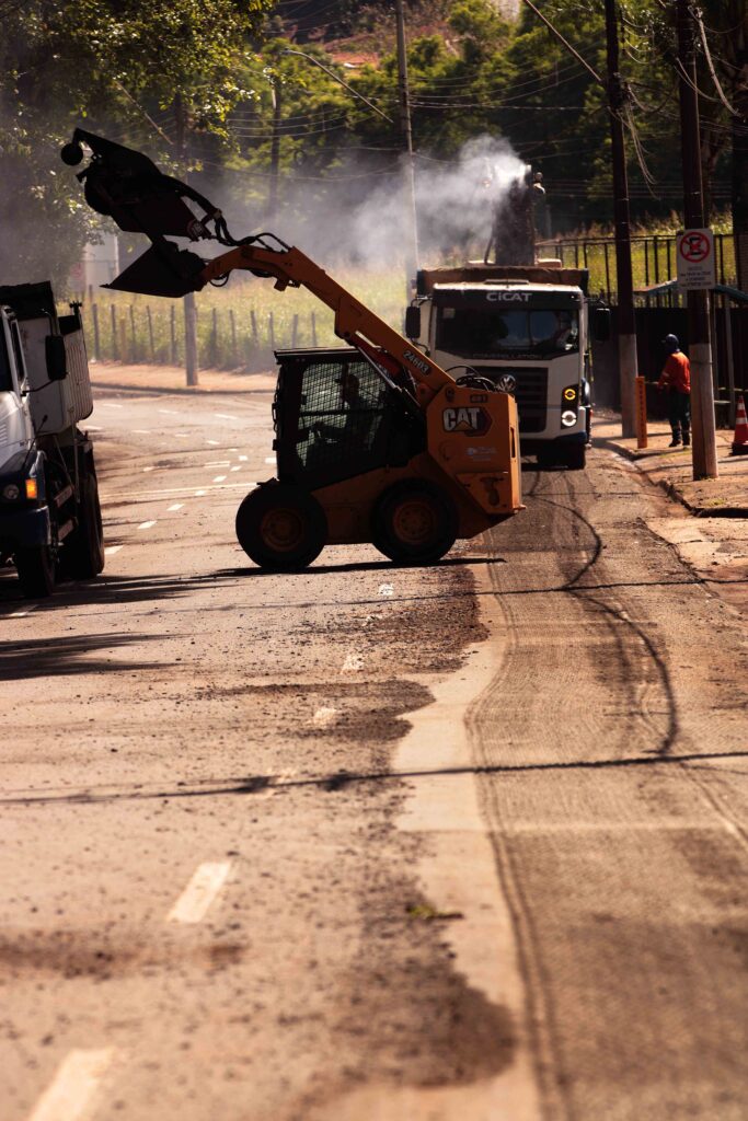 Uma minicarregadeira amarela da marca CAT opera numa via pública com asfalto fresado, rodeada por camiões de obras e fumaça ao fundo.