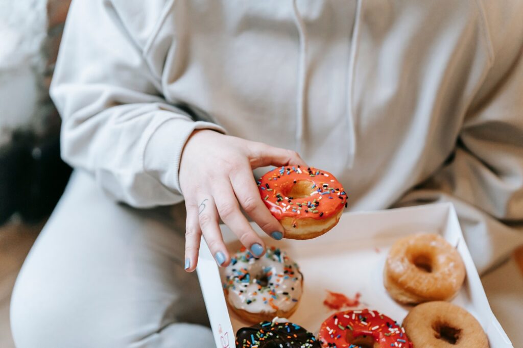 Pessoa a segurar um donut com cobertura cor-de-rosa e granulados coloridos sobre uma caixa de papelão aberta repleta de outros mini donuts de diversos sabores e coberturas.
