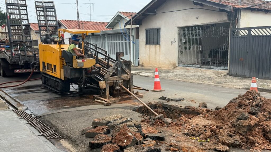 Mostra a máquina perfuratriz amarela operada por um técnico na via pública, realizando a perfuração para a passagem da nova tubulação com cones de sinalização ao redor.