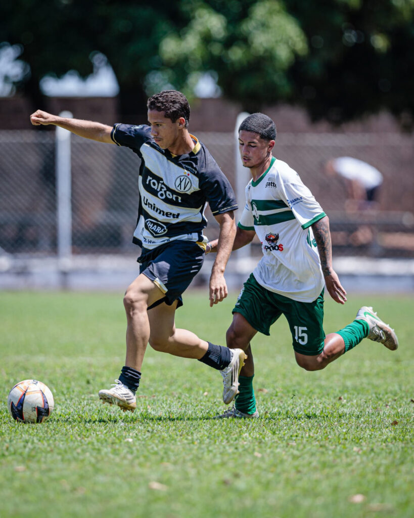 Close-up de ação em um jogo de futebol. Um jogador com o uniforme preto e branco (do XV) dribla a bola com velocidade, sendo perseguido de perto por um adversário com uniforme branco e verde (número 15)