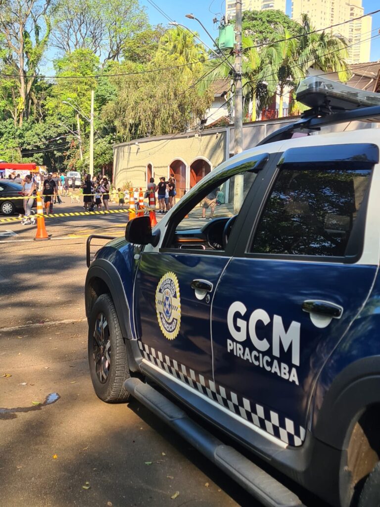 A lateral de um veículo da Guarda Civil Municipal de Piracicaba estacionado numa rua, enquanto, ao fundo, uma área está isolada por fitas e cones para a realização de um evento ao ar livre com público.