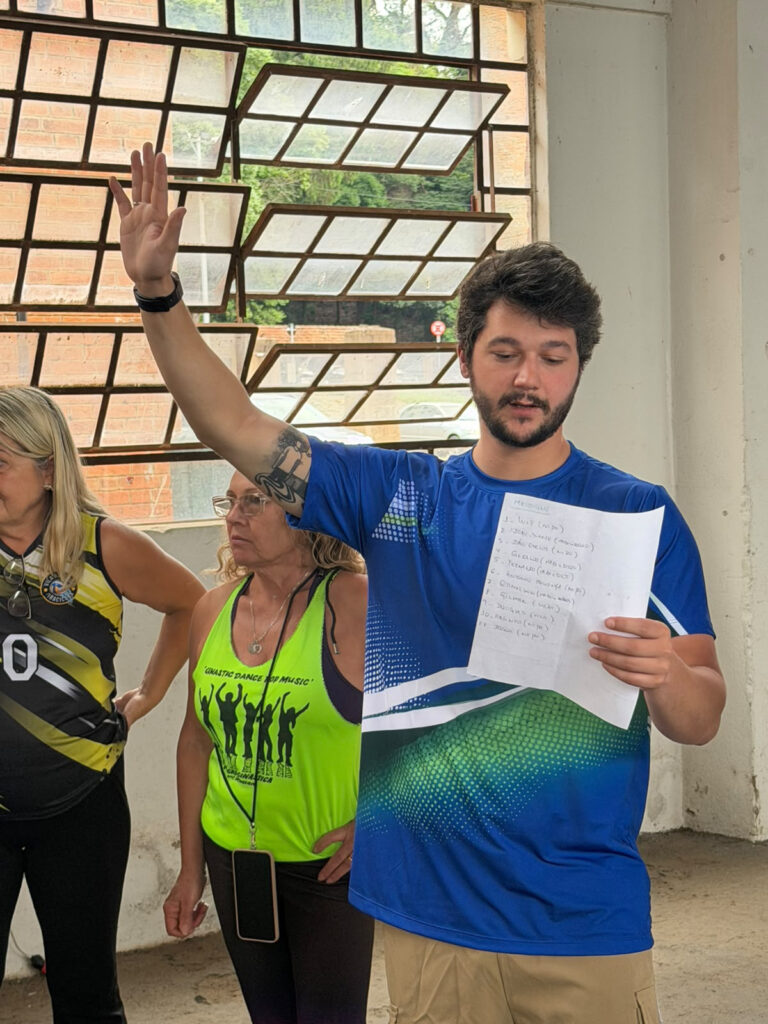 O coordenador da competição surge a ler uma folha de papel com anotações e a erguer o braço direito para orientação dos participantes. Ele veste uma t-shirt desportiva azul e está acompanhado por outros participantes ao seu lado.