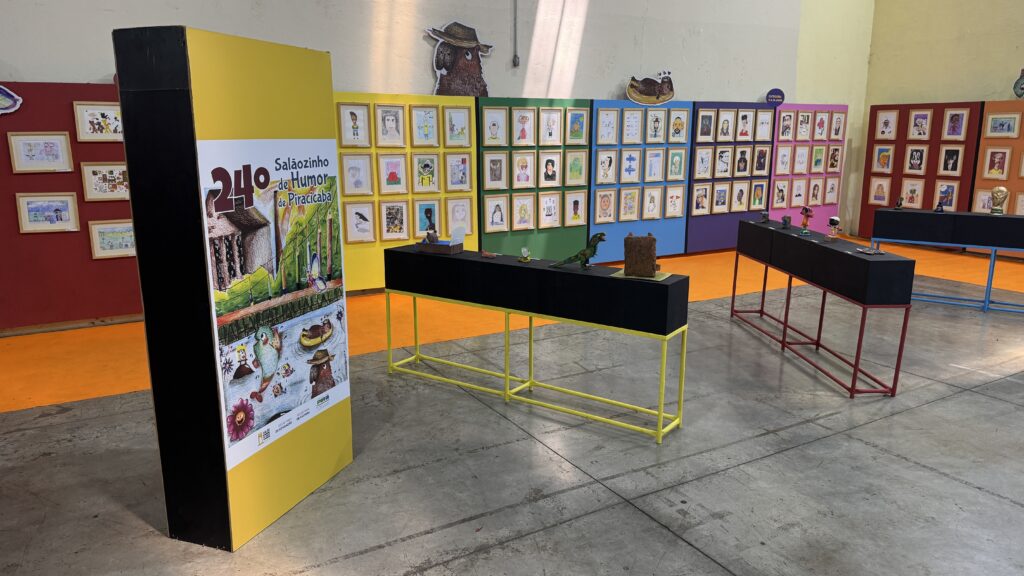 Vista geral da exposição do 24º Salãozinho de Humor de Piracicaba, com painéis coloridos repletos de desenhos emoldurados e bancadas pretas a exibir esculturas infantis.