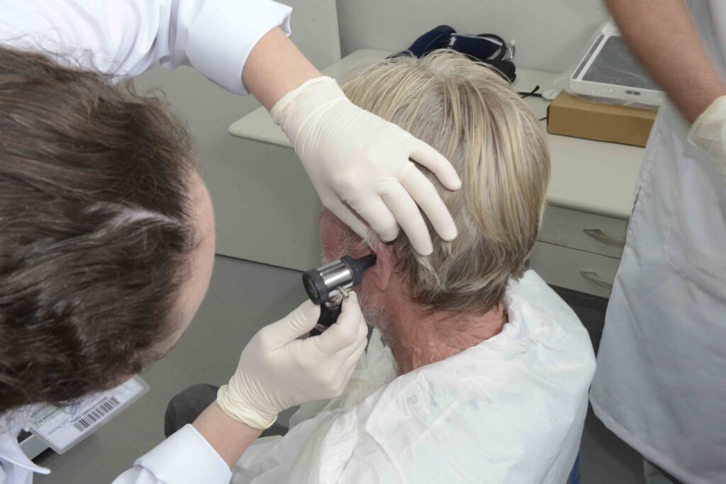 Um profissional de saúde com luvas de proteção utiliza um otoscópio para examinar o ouvido de um paciente sénior.