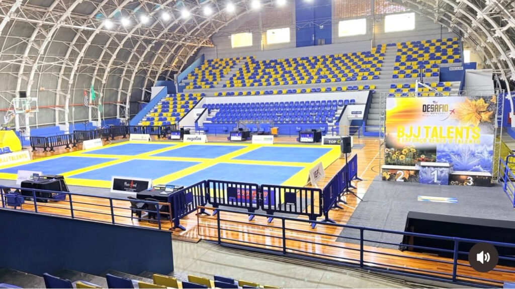 Vista ampla do interior do Ginásio Municipal Waldemar Blatkauskas, mostrando a quadra montada com tatames azuis e amarelos, arquibancadas vazias ao fundo e a estrutura de pódio do evento "BJJ Talents".