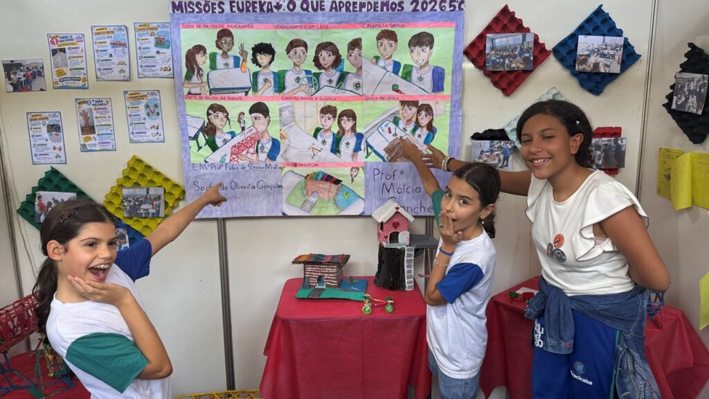Três alunas da rede municipal sorriem e apontam para um cartaz ilustrado de trabalhos escolares intitulado "Missões Eureka"