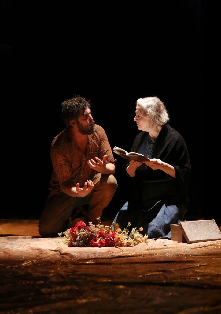 Os atores Iano Salomão e Denise Del Vecchio encenam uma cena dramática ajoelhados sobre um piso revestido, com flores dispostas ao centro, durante a peça Tom na Fazenda