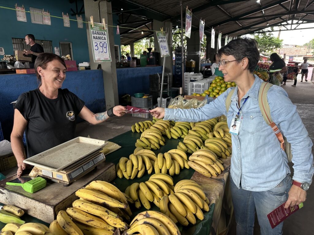 A mesma agente entrega um folheto informativo à feirante Iara Aparecida Stoco, que sorri junto à sua banca de bananas num varejão.