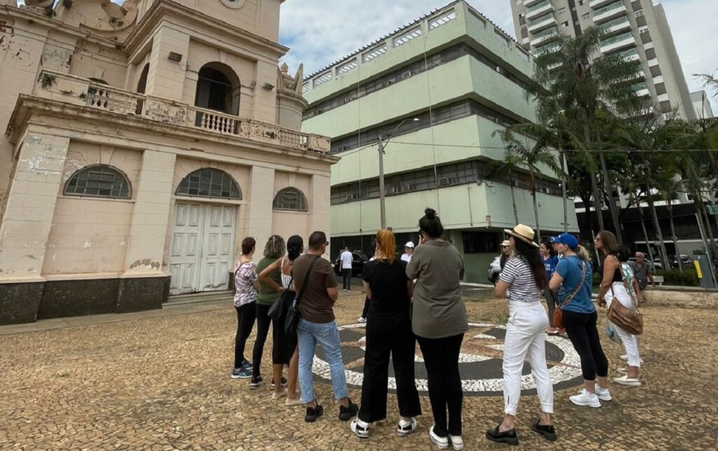Um grupo de pessoas reunido de costas numa praça em frente à Igreja São Benedito, a observar a fachada do edifício histórico e os prédios ao redor.