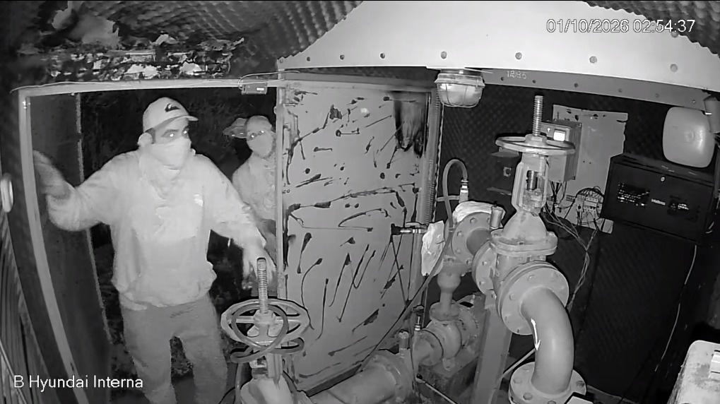 Imagem preto e branco de câmara de segurança interna, mostrando dois homens com o rosto parcialmente coberto (máscaras/tecidos) junto a estruturas metálicas, válvulas e tubagens de uma estação.