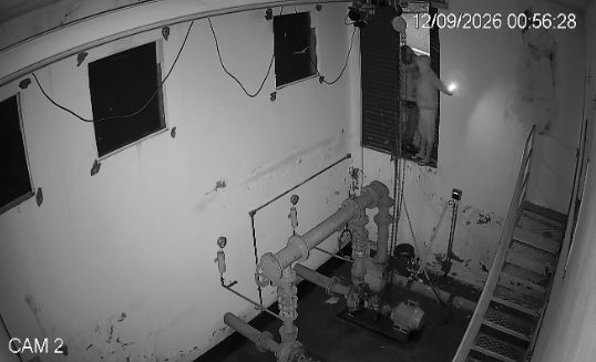 Imagem a preto e branco captada por câmara de segurança (CAM 2), mostrando o interior de uma instalação com tubagens e escadas, onde dois indivíduos com lanterna parecem entrar ou espreitar através de uma janela elevada.