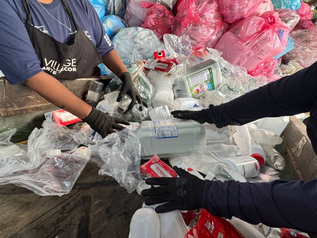 Trabalhadores da cooperativa de reciclagem, utilizando luvas de proteção, realizam a triagem manual de resíduos plásticos e embalagens numa esteira de separação.