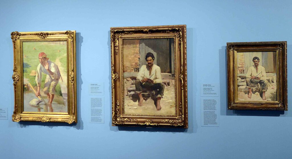 Três quadros de Almeida Júnior expostos lado a lado numa parede azul com etiquetas explicativas, incluindo estudos e obras da exposição Caipiras.