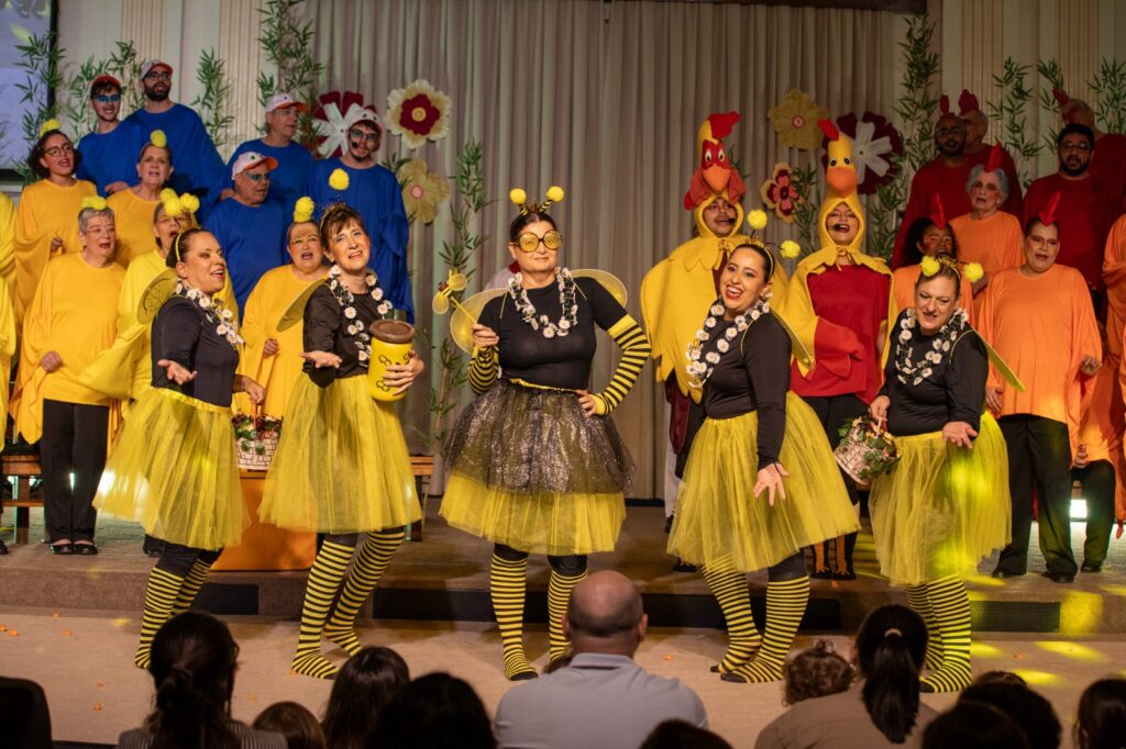 Atores vestidos de abelhas e galinhas apresentam-se na frente do palco, acompanhados ao fundo por um grupo de coralistas com túnicas coloridas no Teatro Erotides de Campos.