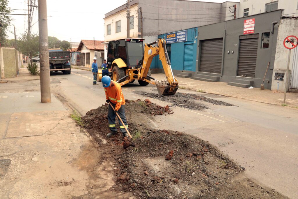 Um trabalhador em primeiro plano utiliza uma pá para nivelar montes de terra e detritos na via pública, enquanto uma retroescavadora e outros operários dão continuidade às obras de requalificação urbana ao fundo.
