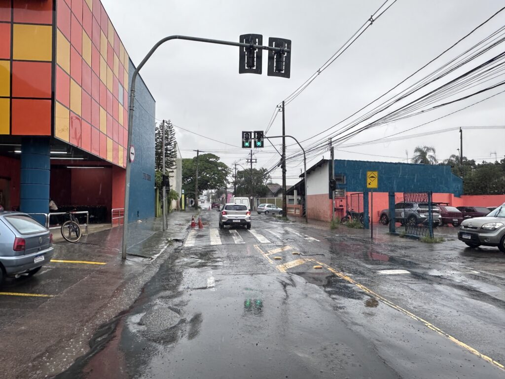 A imagem mostra uma rua urbana molhada pela chuva, vista a partir da perspetiva de um condutor num dia nublado. Do lado esquerdo, destaca-se um edifício comercial com fachada colorida em quadrados amarelos, laranjas e vermelhos. Ao centro, vê-se um cruzamento com semáforos, uma passagem de peões e alguns carros em circulação numa via de asfalto desgastado. O ambiente reflete condições de tempo chuvoso.