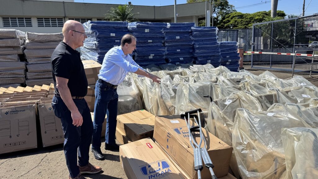 Dois homens observam e inspecionam o lote de doações no pátio. Em primeiro plano sobressaem caixas de papelão, muletas de alumínio, pilhas de colchões empilhados e dezenas de cadeiras de rodas e equipamentos envolvidos em plástico transparente.