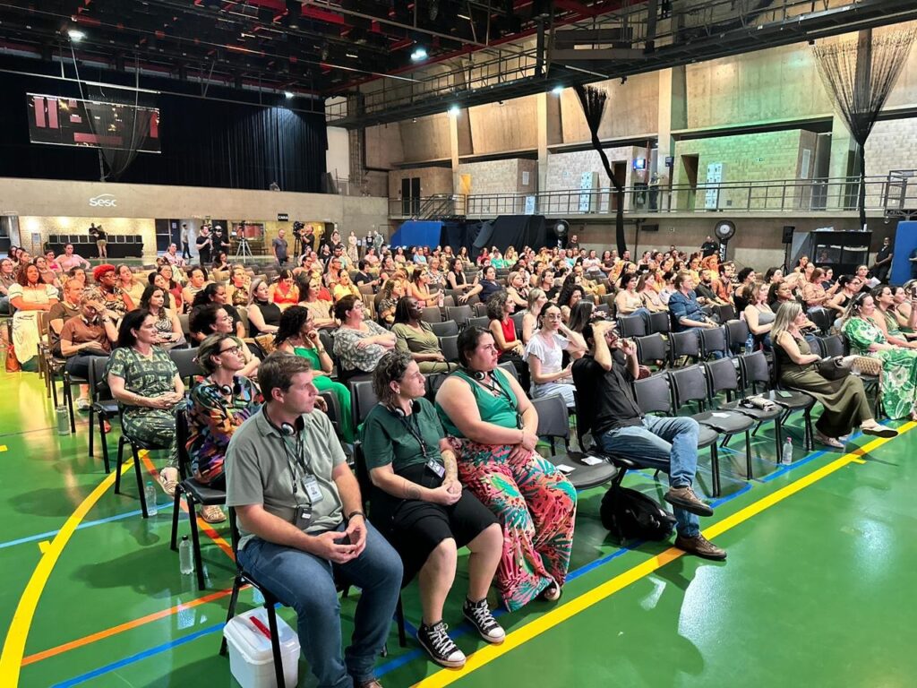 Vista ampla do público acomodado em cadeiras na quadra coberta do Sesc durante a edição da Jornada Pedagógica.