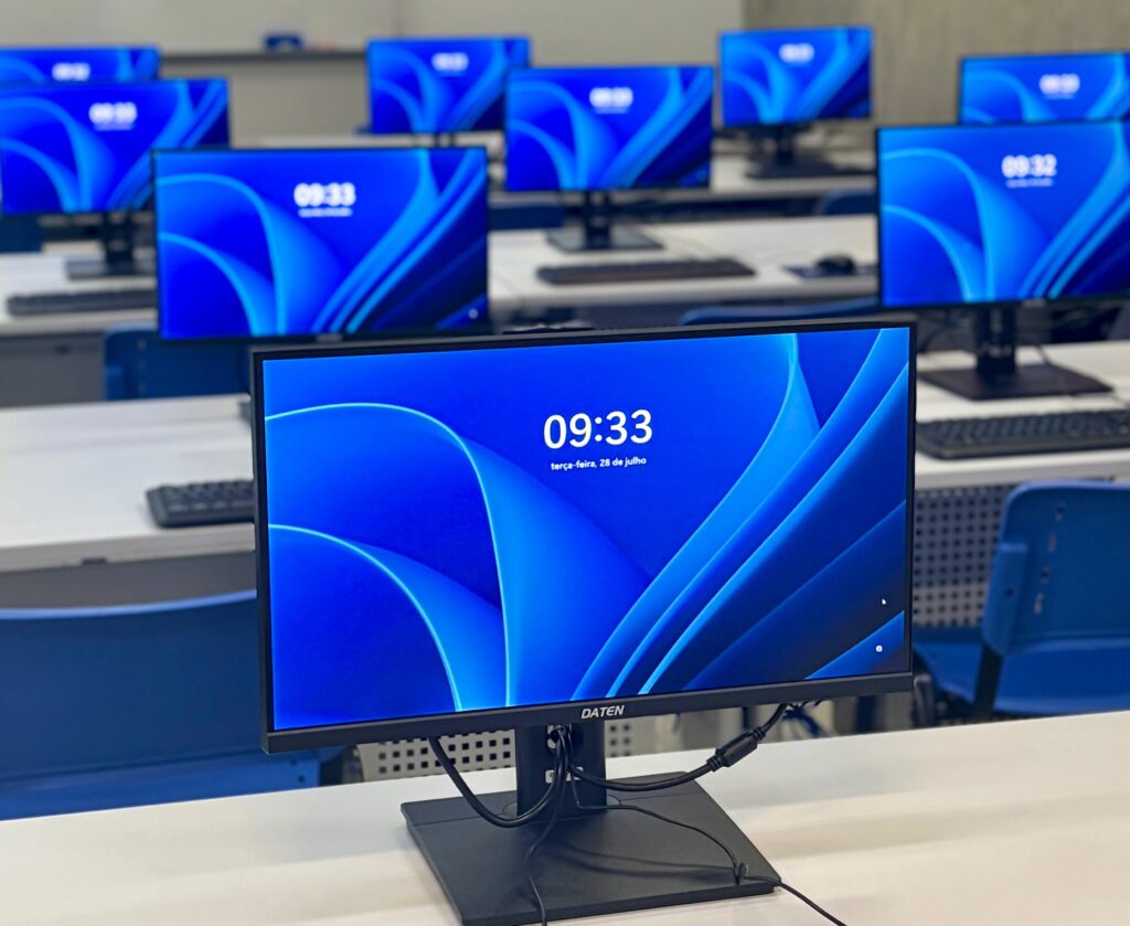 Vista em detalhe dos novos computadores da marca DATEN alinhados sobre as bancadas brancas do laboratório, com ecrãs azuis acesos a exibir a hora e a data.