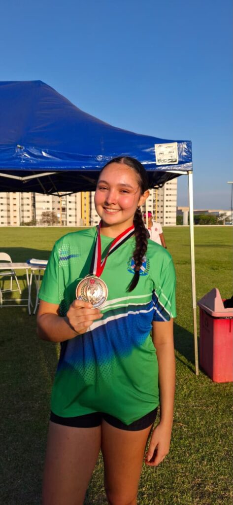 Jovem atleta em pé ao ar livre, vestindo o uniforme desportivo verde e azul de Piracicaba e a segurar a medalha de bronze conquistada no salto em distância.