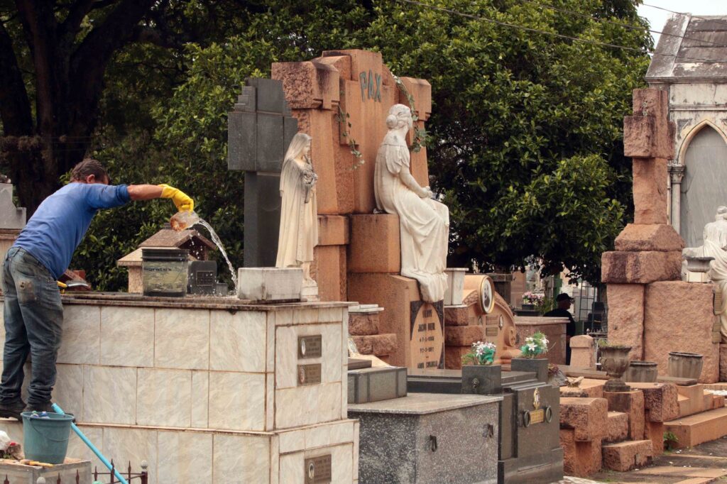 Mostra uma pessoa de luvas amarelas a lavar e a deitar água sobre a estrutura de um jazigo num cemitério, rodeada por baldes e com várias estátuas e túmulos ornamentados ao fundo.