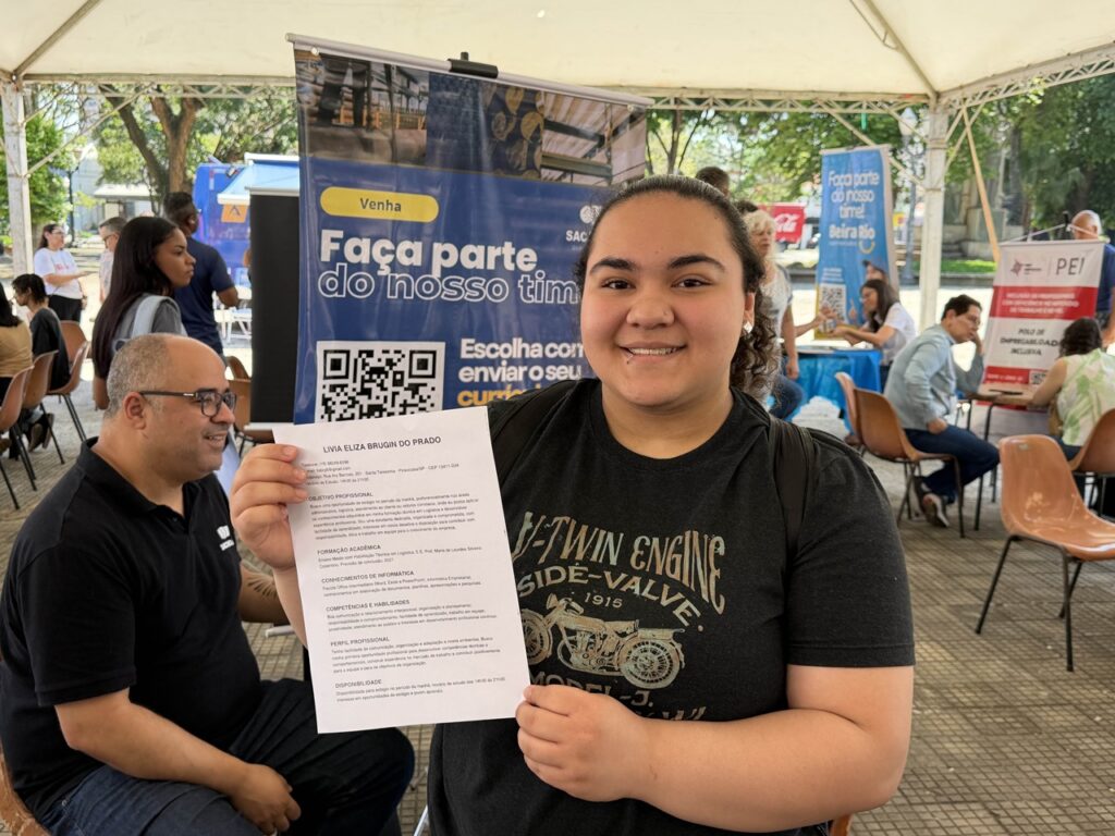 A jovem Lívia Eliza Brugin do Prado sorri em primeiro plano enquanto segura e exibe o seu currículo impresso no local do evento.