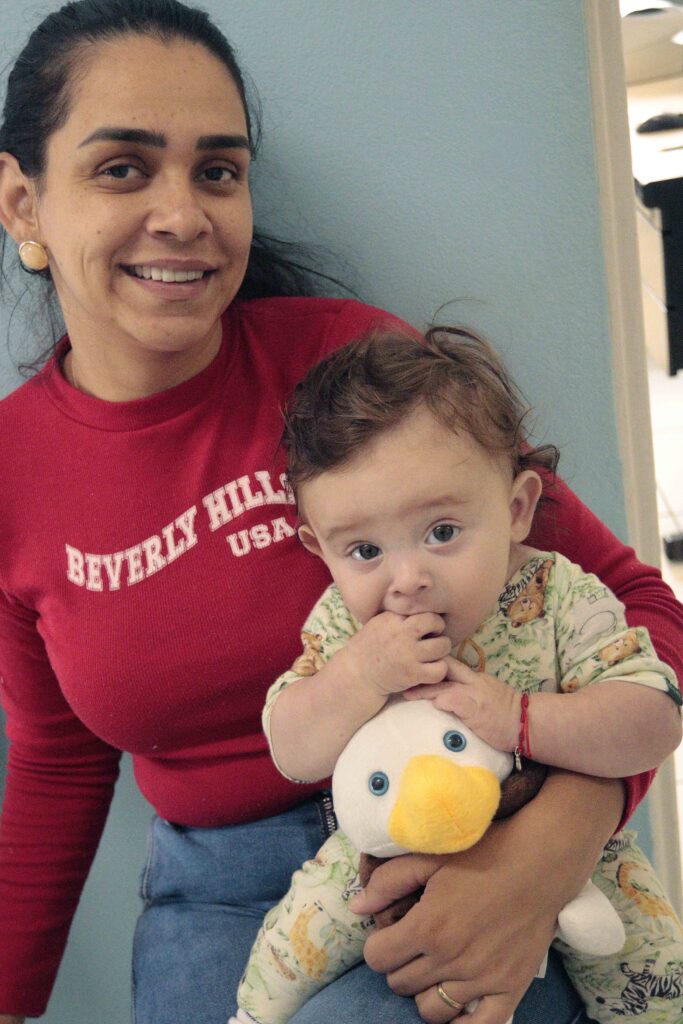 Luana Iaccope posa sorridente com o seu filho bebé, Diogo, ao colo, que segura e morde um peluche de pato doado pela agente de saúde.