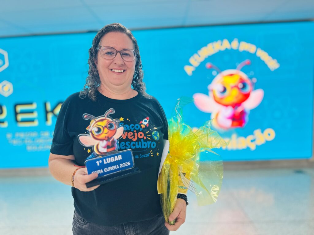 Uma mulher de óculos e cabelo encaracolado sorri, exibindo um troféu de 1.º lugar da Feira Eureka 2026 numa mão e um presente embrulhado em celofane amarelo na outra.