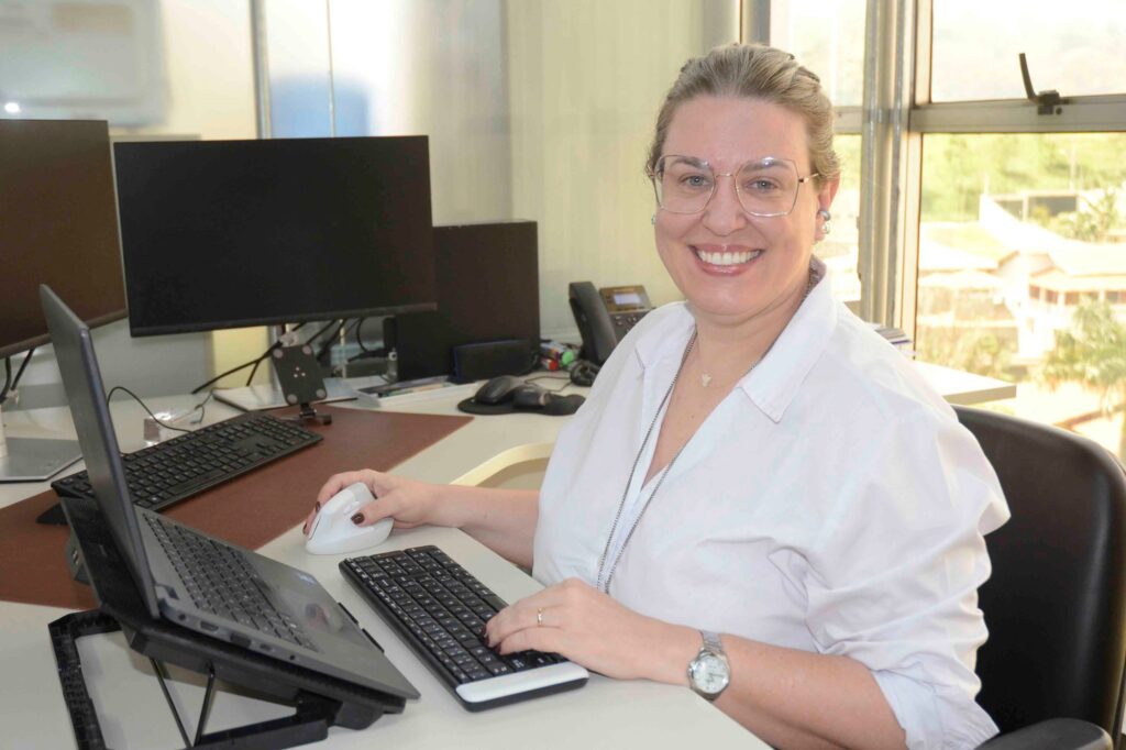 Luciane Leães sorri para a câmara sentada à sua secretária de trabalho num escritório, enquanto utiliza o portátil e o computador.