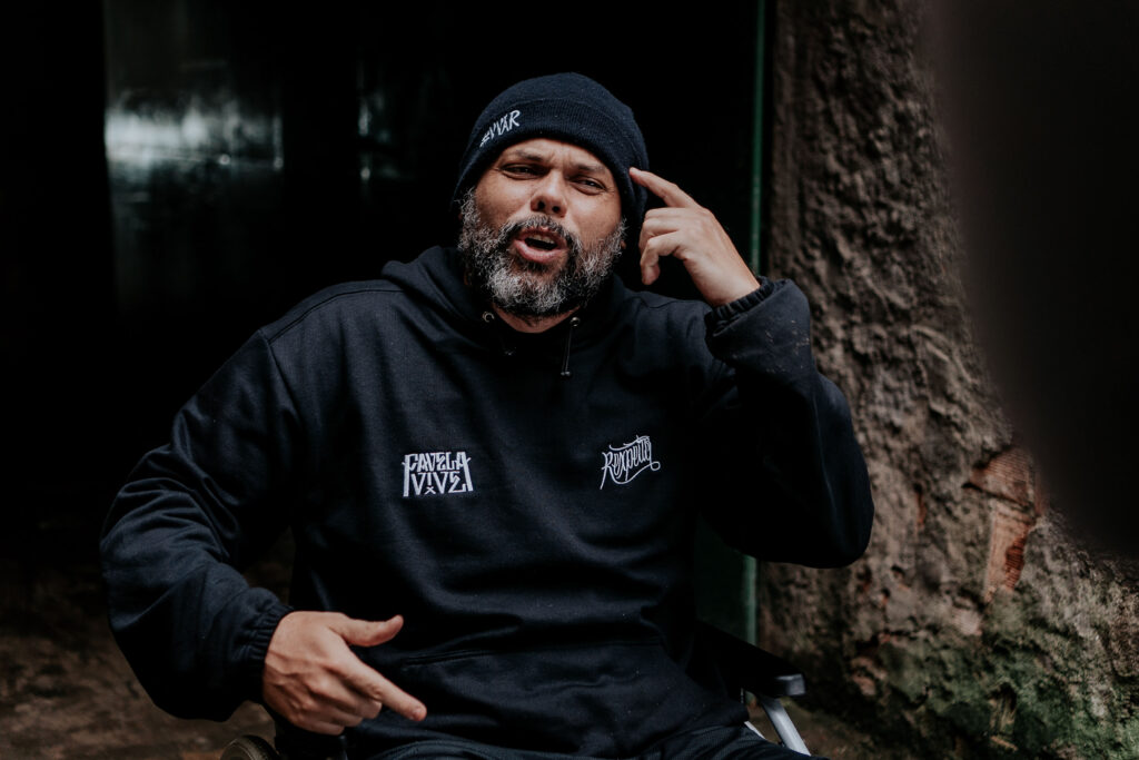 O rapper MC Marechal gesticula com a mão na cabeça enquanto fala, usando gorro preto e casaco escuro de capuz.