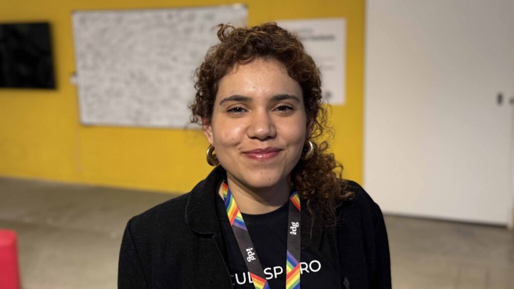 Retrato em plano médio da orientadora pedagógica Maiana Monteiro, sorridente, a utilizar um fita de pescoço do programa de formação cultural sobre um fundo amarelo.