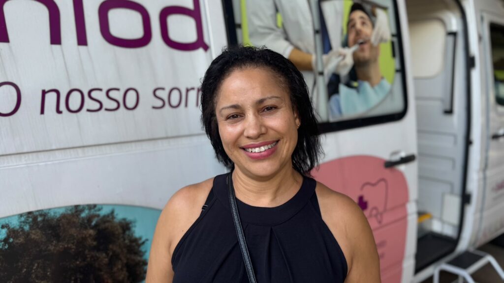Uma mulher sorri para a câmara num plano aproximado, posicionada em frente a uma unidade móvel de saúde (odontomóvel).